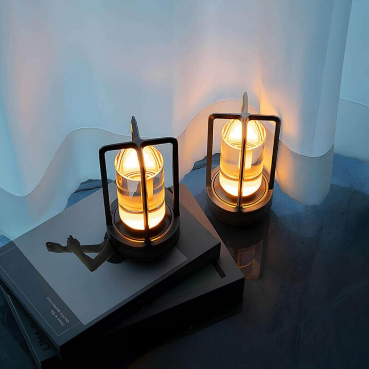 Dvě svítící lampy s tmavým rámem a skleněným válcem stojí na tmavých knihách před průsvitnou bílou záclonou. 3