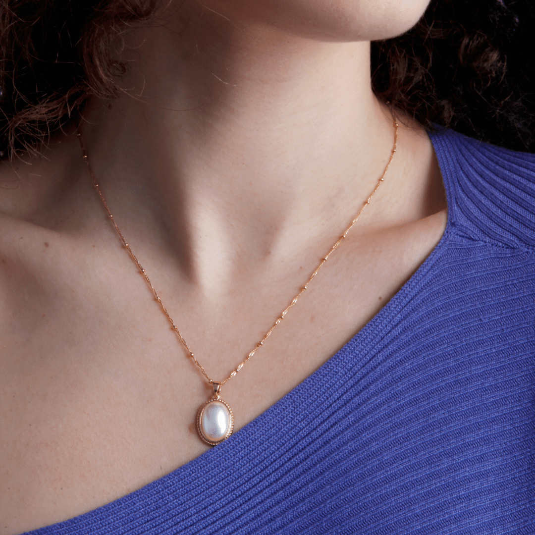 Frau mit goldener Halskette und Perlenanhänger, blaues Oberteil, eleganter Schmuck.