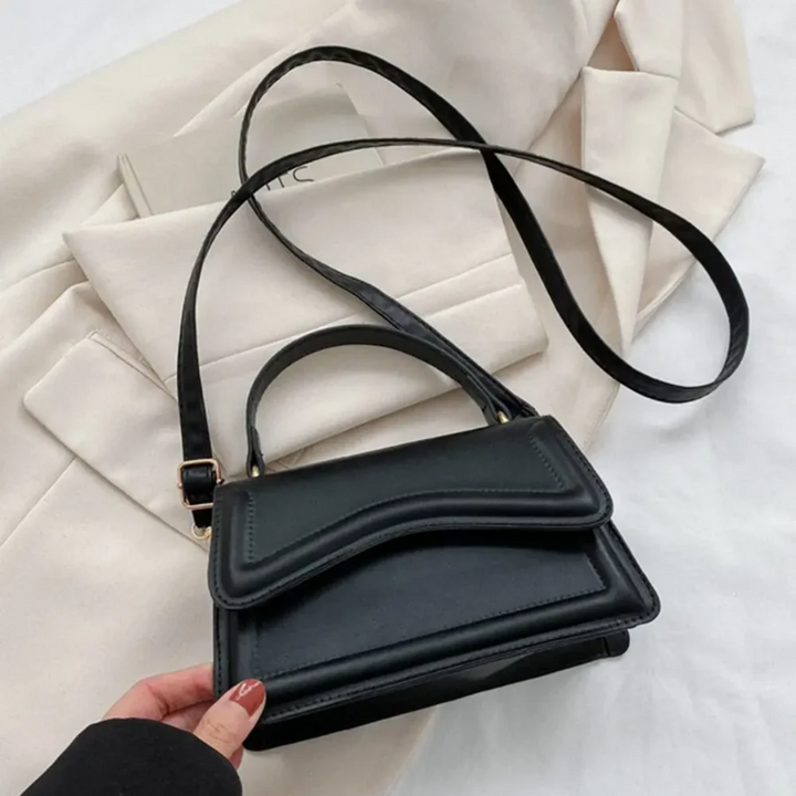 Schwarze Lederhandtasche mit verstellbarem Riemen auf beigem Stoffhintergrund.