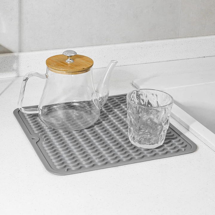 Glas-Teekanne mit Holzdeckel und Glas auf grauer Silikon-Abtropfmatte in moderner Küche.