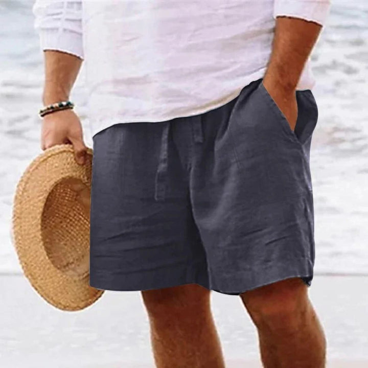 Mann in weißem Hemd und dunkelblauer Leinen-Shorts am Strand, hält Strohhut.