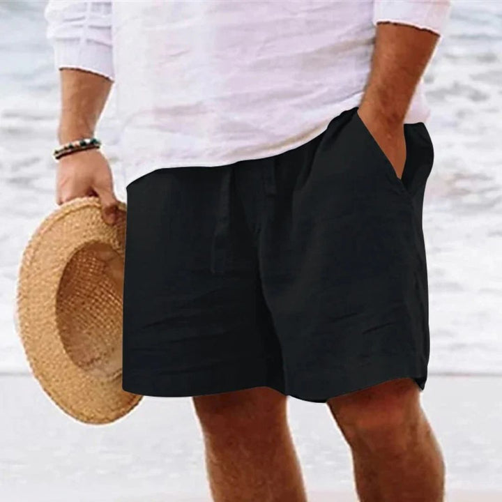 Mann in schwarzer Leinen-Shorts und weißem Hemd am Strand, hält Strohhut in der Hand.