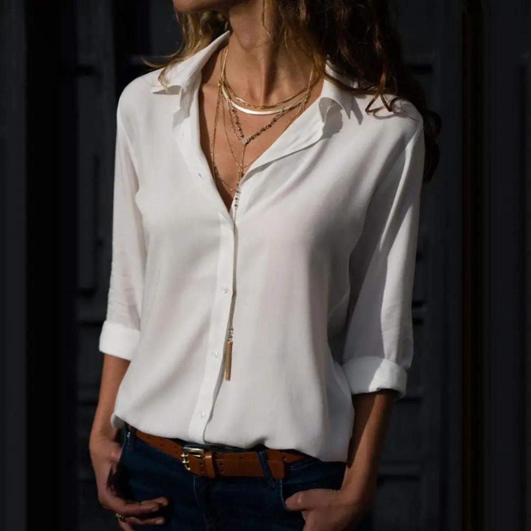 Frau in weißer Bluse, brauner Gürtel, goldene Halsketten, modisches Outfit, dunkler Hintergrund.