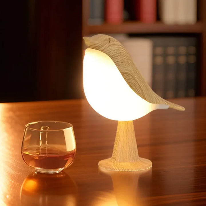 Holz-Vogel-Tischlampe mit warmem Licht neben einem Glas Whisky auf Holztisch.