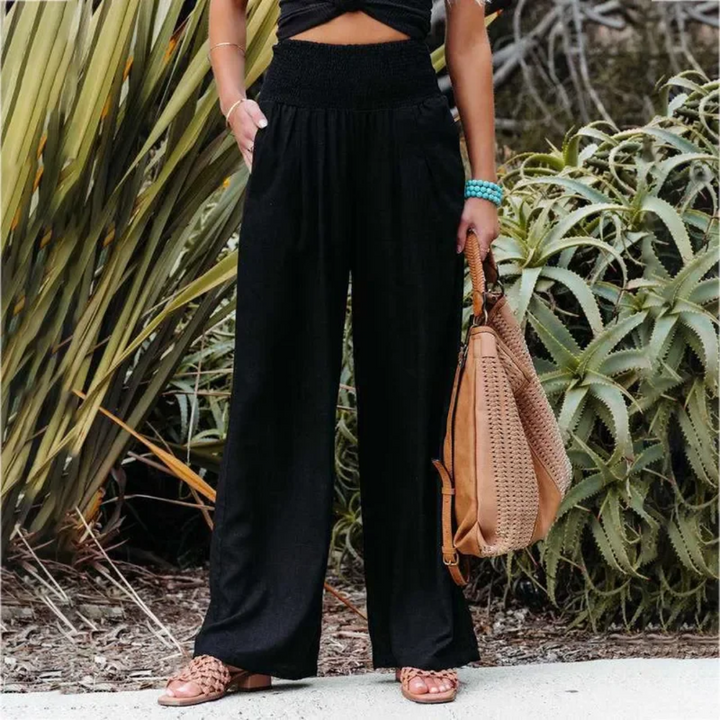 Frau in schwarzer Hose, hält braune Tasche, steht vor Pflanzen, Sommermode, Freizeitlook.
