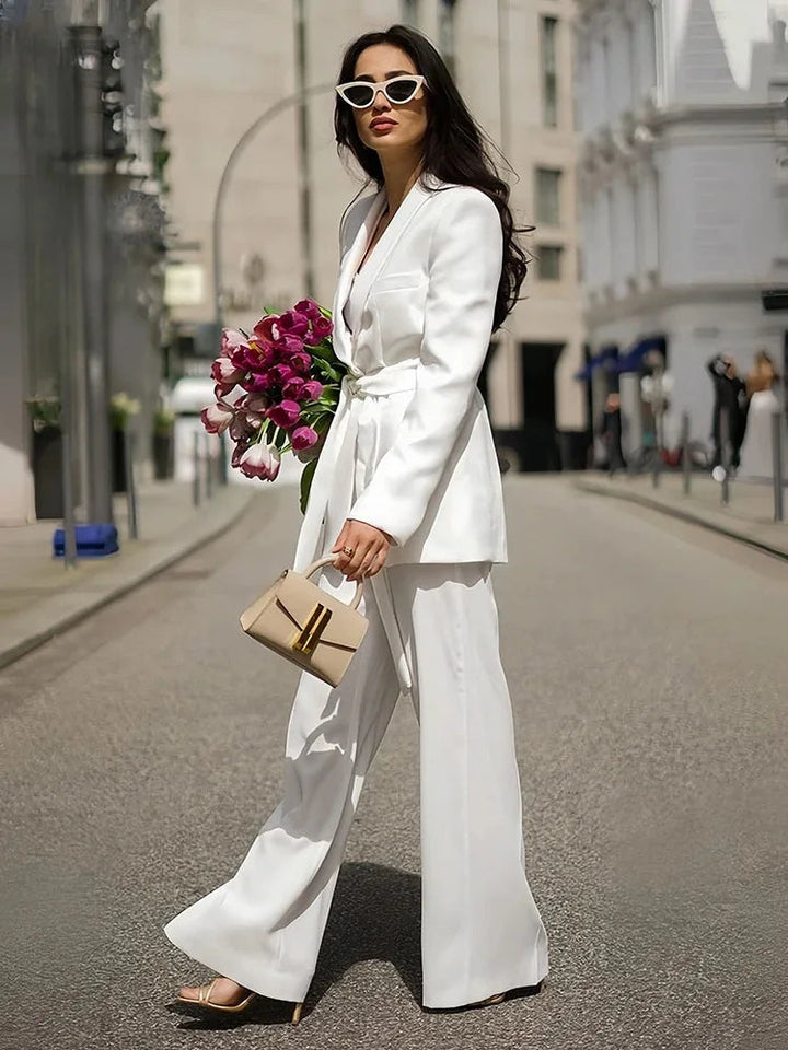 Frau in weißem Anzug mit Gürtel, Sonnenbrille, Blumenstrauß und Handtasche auf Stadtstraße.