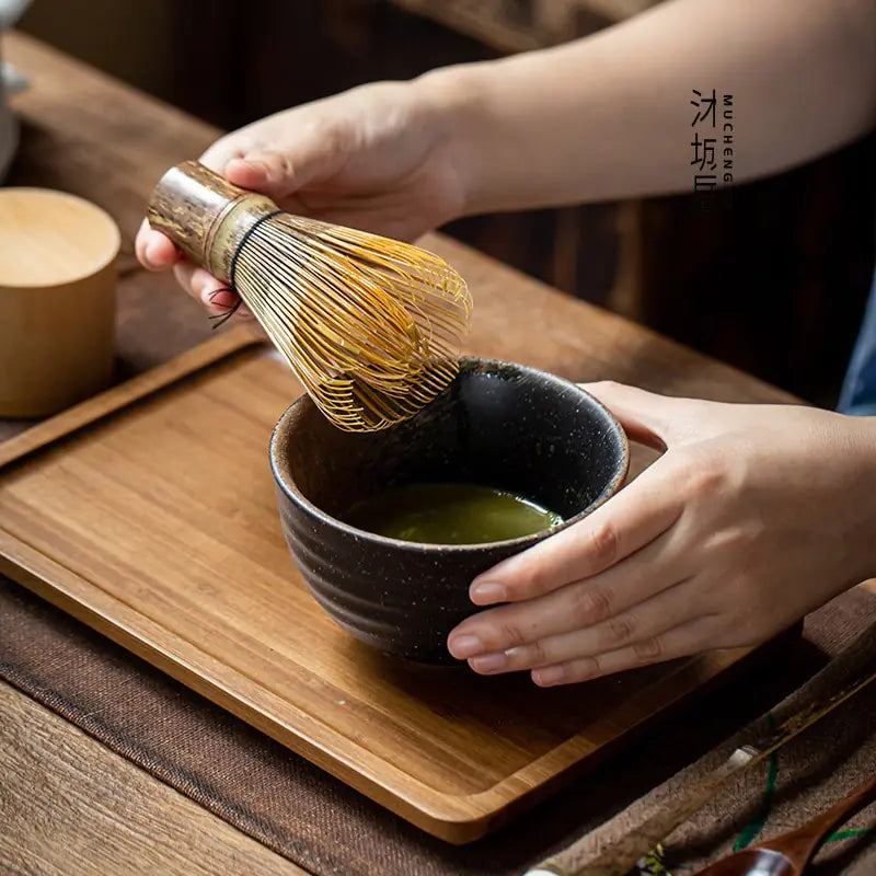Hand hält Bambus-Teebesen über schwarzer Schale auf Holztablett, Matcha-Zubereitung, japanisch.