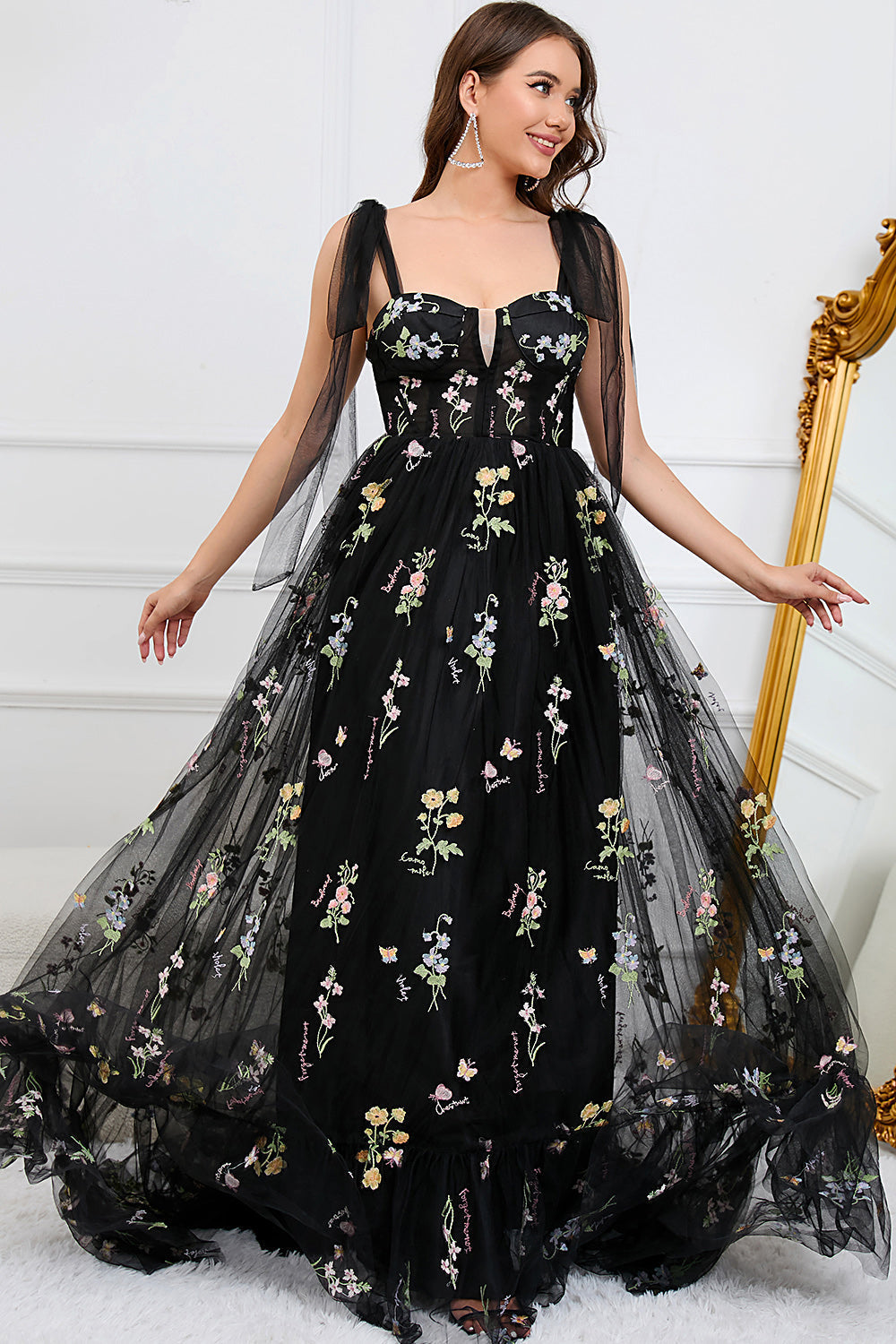 Frau in schwarzem Abendkleid mit Blumenstickerei vor einem goldenen Spiegel.