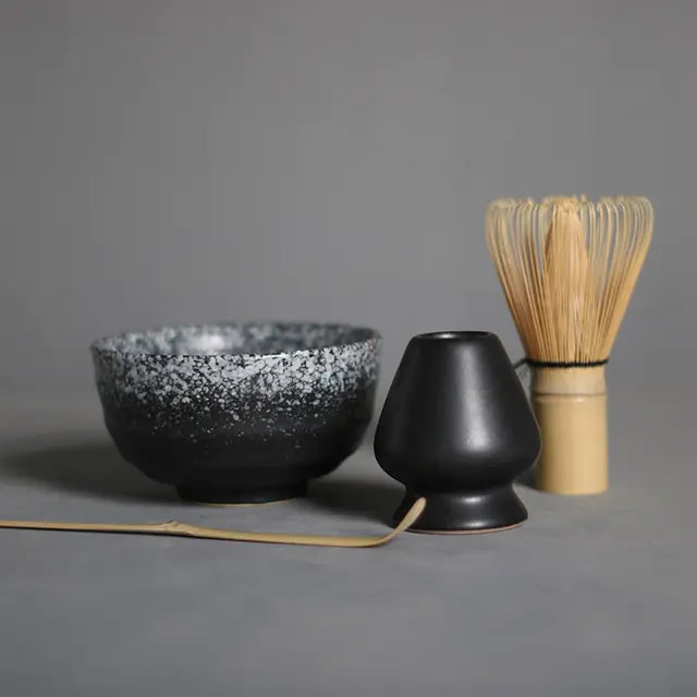 Schwarze Keramik-Teeschale, Bambusbesen und Löffel für Matcha-Zubereitung auf grauem Hintergrund.