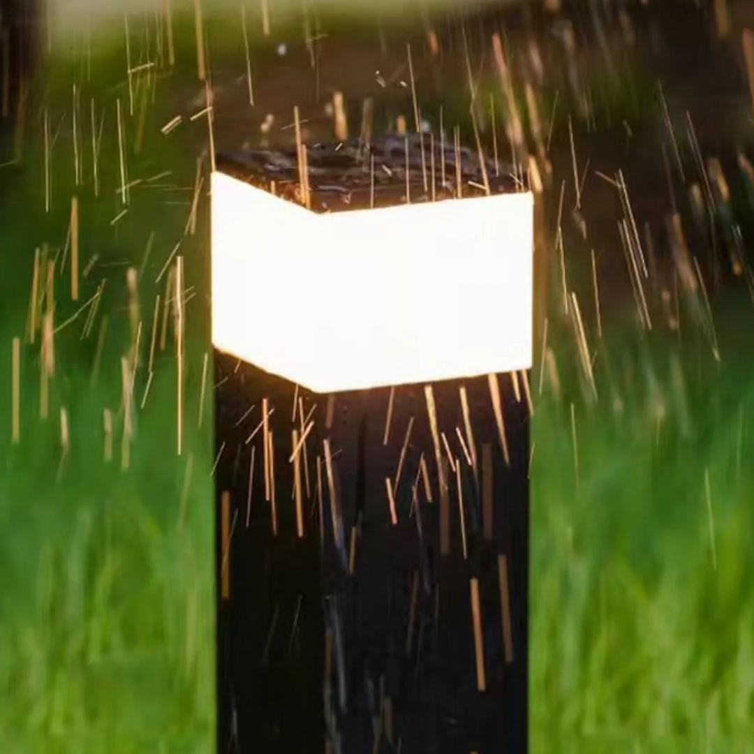 Detail horní části svítícího sloupku v dešti, kapky vody dopadají na tmavé víko a kryt. 9