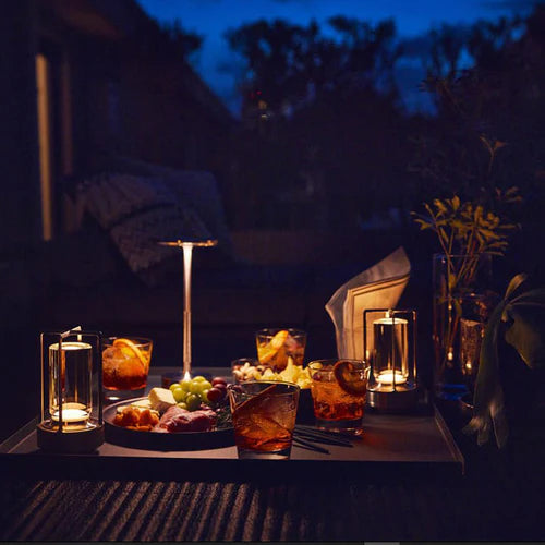 Moderne Tischleuchte bei Nacht, umgeben von Getränken und Snacks auf einem Tisch im Freien.