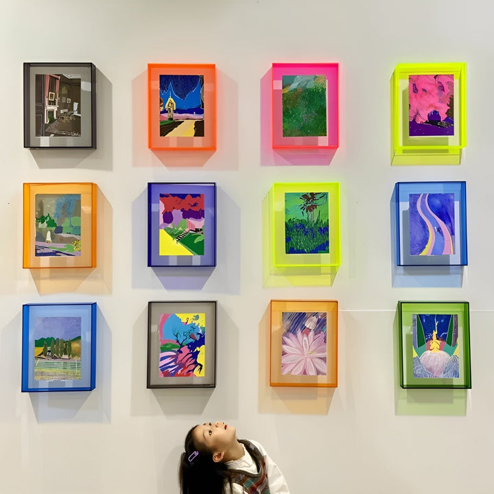 Nástěnná galerie dvanácti barevných akrylových rámečků v neonových odstínech s malbami, pod nimi se dívá vzhůru malá holčička. 22