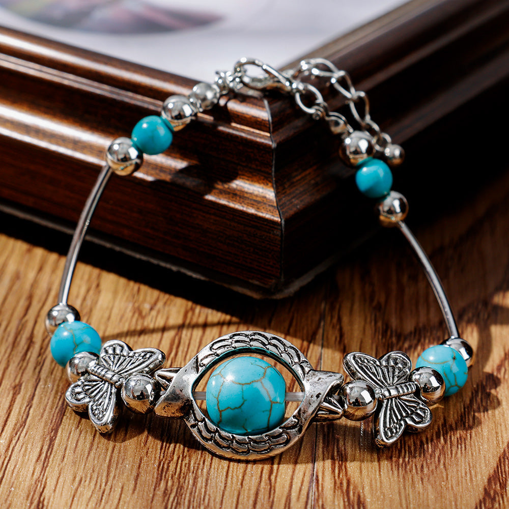 Silberarmband mit türkisfarbenen Steinen und Schmetterlingsanhängern auf Holzhintergrund.
