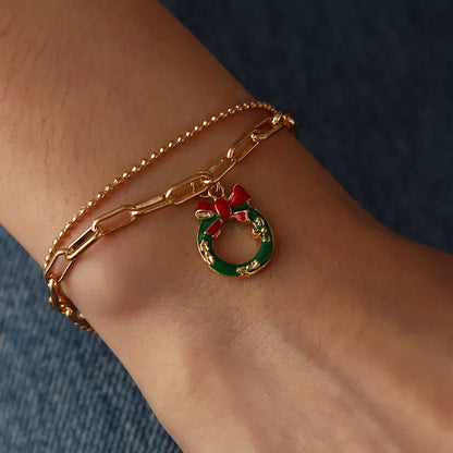 Goldenes Armband mit grünem Weihnachtskranz-Anhänger und roter Schleife auf Handgelenk.