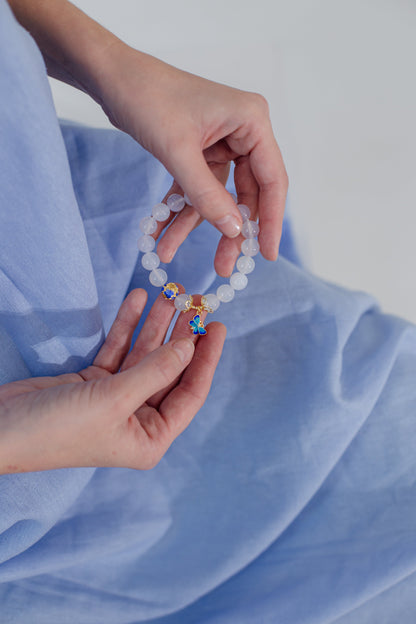 Hände halten weißes Perlenarmband mit bunten Anhängern, blauer Stoffhintergrund.
