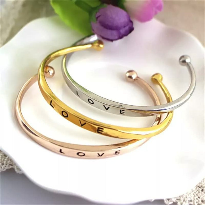Drei Armreifen aus Metall in Gold, Silber und Roségold mit Gravur "LOVE" auf weißem Teller.