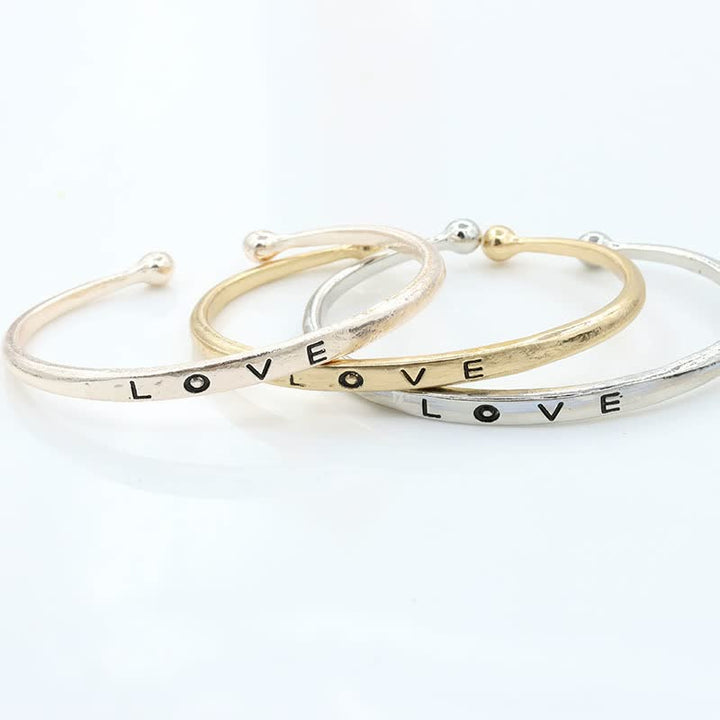Drei Armreifen aus Metall in Gold, Silber und Roségold mit "LOVE"-Gravur auf weißem Hintergrund.