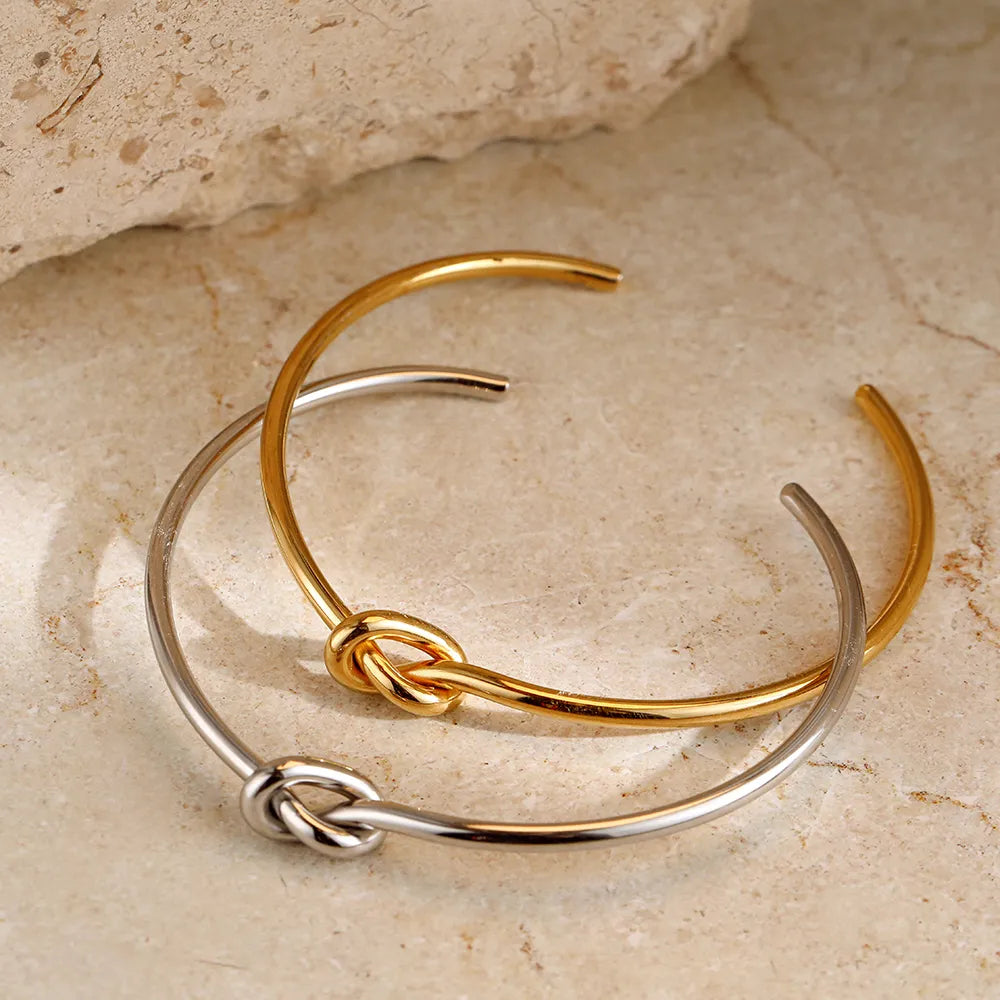 Zwei offene Armreife aus Gold und Silber mit Knoten-Design auf beigem Steinuntergrund.