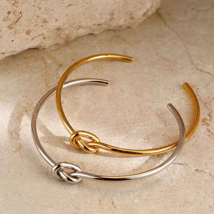 Zwei offene Armreife aus Gold und Silber mit Knoten-Design auf beigem Steinuntergrund.