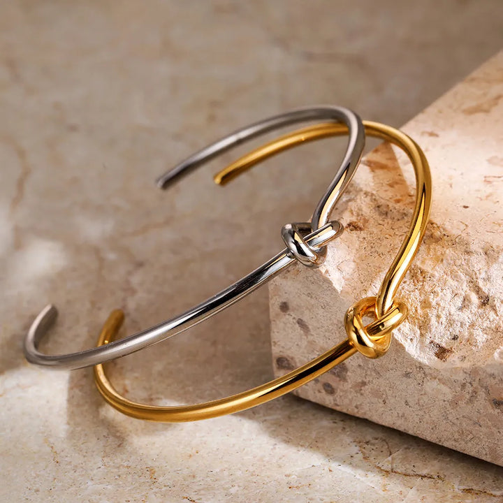 Zwei offene Armreifen aus Edelstahl, gold und silber, mit Knoten-Design auf Steinuntergrund.