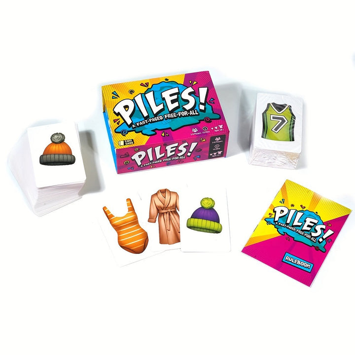 Kartenspiel "PILES!" mit Kartenstapel, Regelbuch und bunter Verpackung auf weißem Hintergrund.