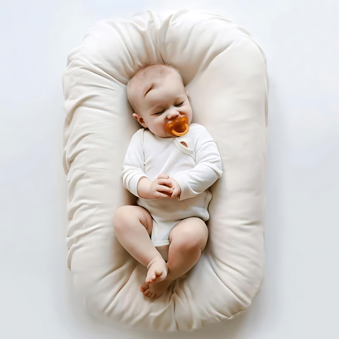 Schlafendes Baby in weißem Strampler auf cremefarbenem Babynest mit Schnuller.