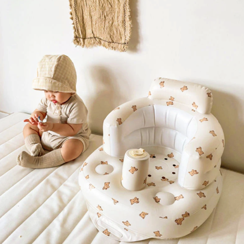 Kleinkind in beigem Outfit neben aufblasbarem Baby-Sitz mit Bärenmuster auf weißer Matratze.