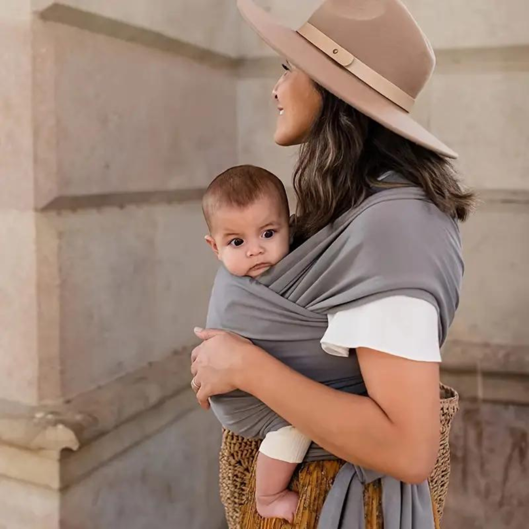 Frau mit Baby in grauem Tragetuch, beige Hut, lächelnd, draußen, Mutter-Kind-Bindung.