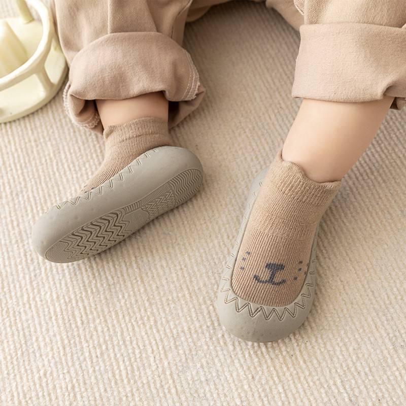 Kleinkind trägt beige rutschfeste Babyschuhe mit Tiermotiv auf Teppich.