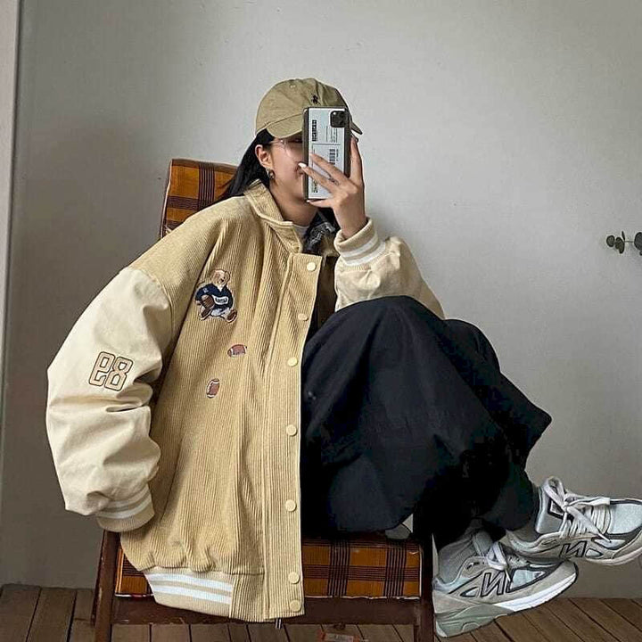 Frau in beige Collegejacke und Kappe, sitzt auf Stuhl, hält Smartphone, modisch, lässig.
