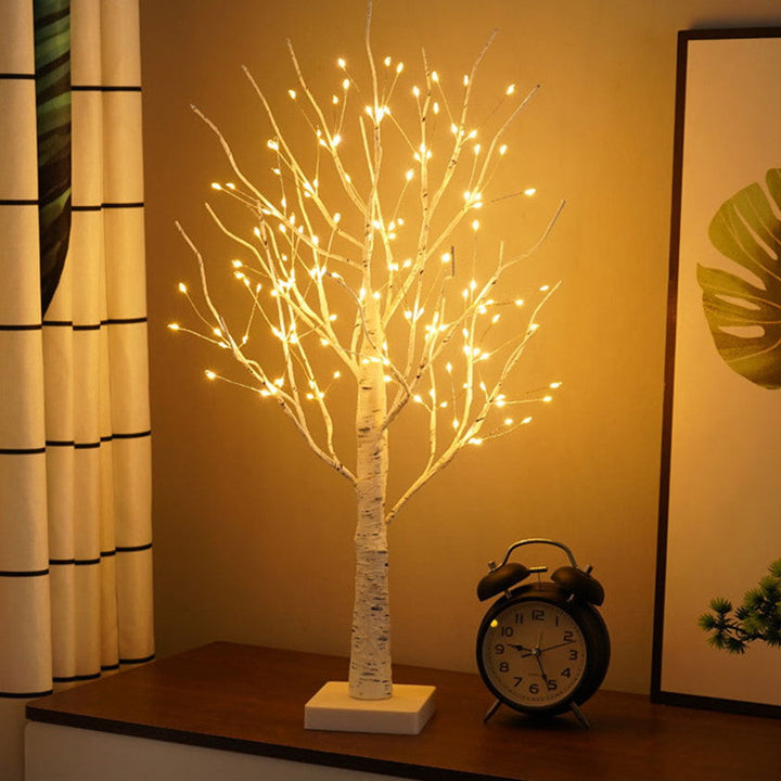 LED-Baumlampe mit warmweißen Lichtern, dekorativ, auf Tisch neben Wecker und Vorhang.