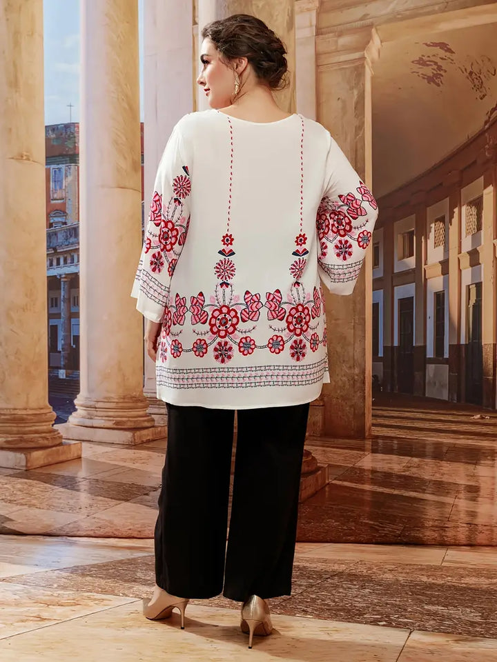 Frau in weißer Tunika mit roten Blumenmustern, schwarze Hose, elegante Umgebung.