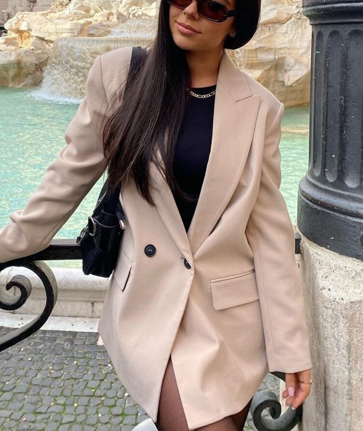 Frau in beigem Blazer, schwarzem Top, Sonnenbrille, vor Brunnen stehend.
