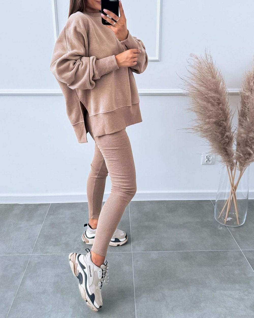Frau in beigem Sweatshirt und Leggings, trägt Sneaker, steht neben Pampasgras, moderne Mode.