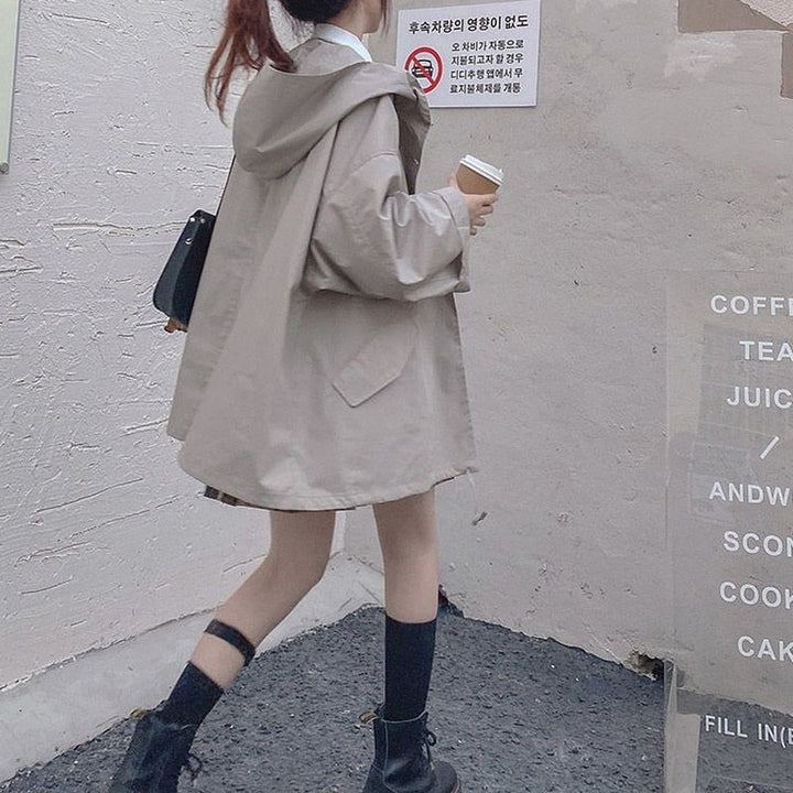 Frau in beigem Oversize-Mantel mit Kapuze, hält Kaffeebecher, trägt schwarze Stiefel.