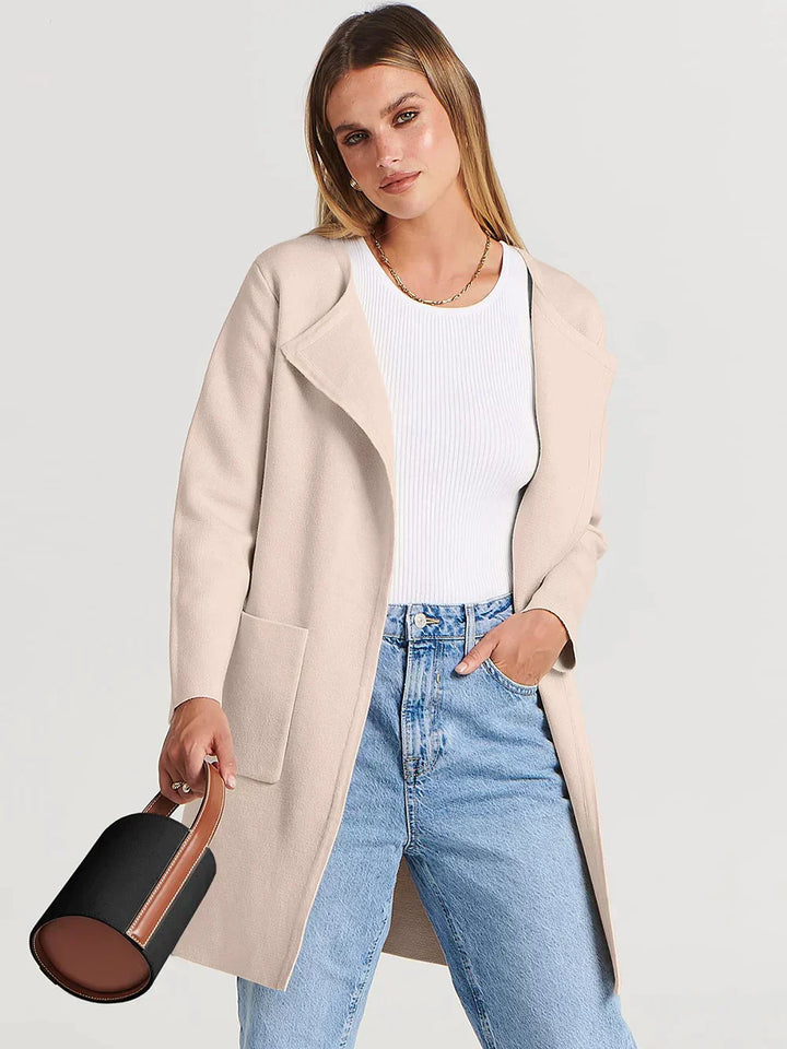 Frau in beigem Wollmantel, weißem Pullover, blauer Jeans, hält schwarze Handtasche.