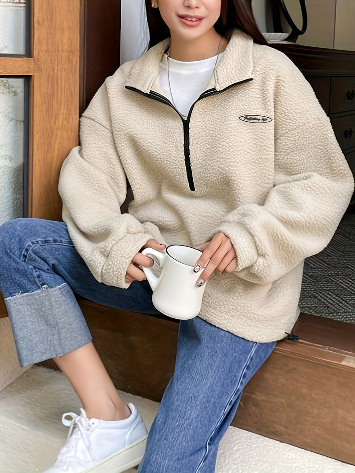 Frau in beiger Fleecejacke mit Reißverschluss, hält weiße Tasse, lässige Jeans und Sneaker.