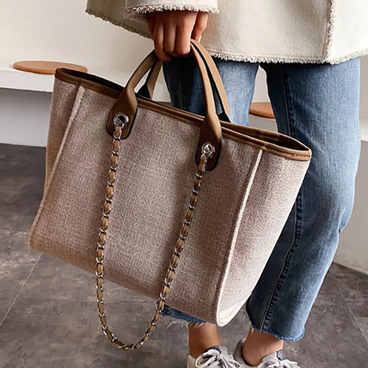 Frau mit beiger Stofftasche, braune Ledergriffe, Kettendetail, lässiger Stil, Jeans, Sneaker.