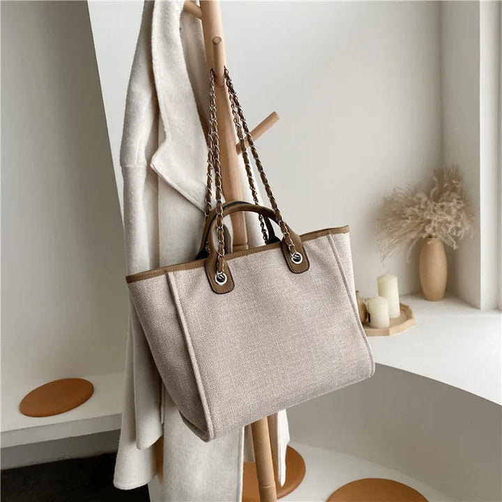 Beige Damenhandtasche aus Leinen mit braunen Ledergriffen, hängend an Holzständer.