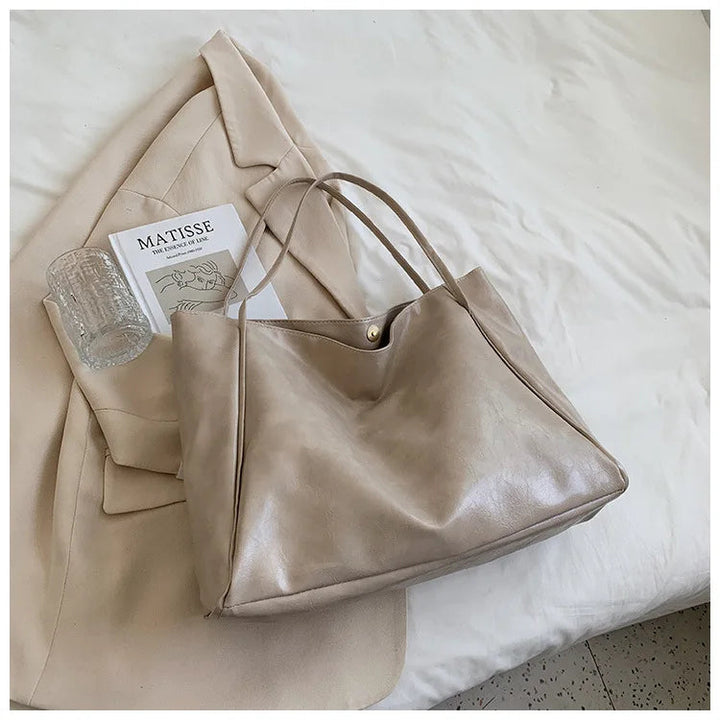 Beige Ledertasche auf beigem Mantel, neben Matisse-Buch und Glas, auf weißem Bett.