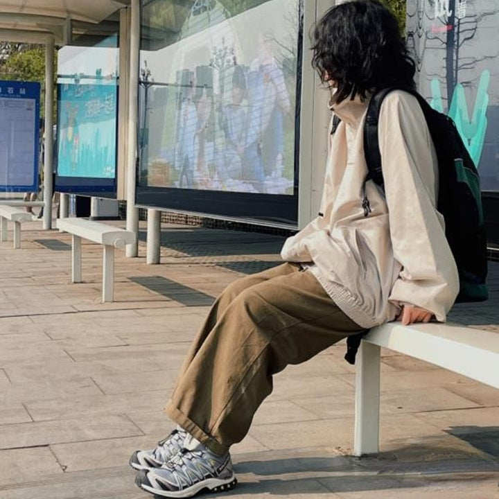 Person in beiger Jacke und brauner Hose sitzt an Bushaltestelle, trägt graue Sneakers, Rucksack.