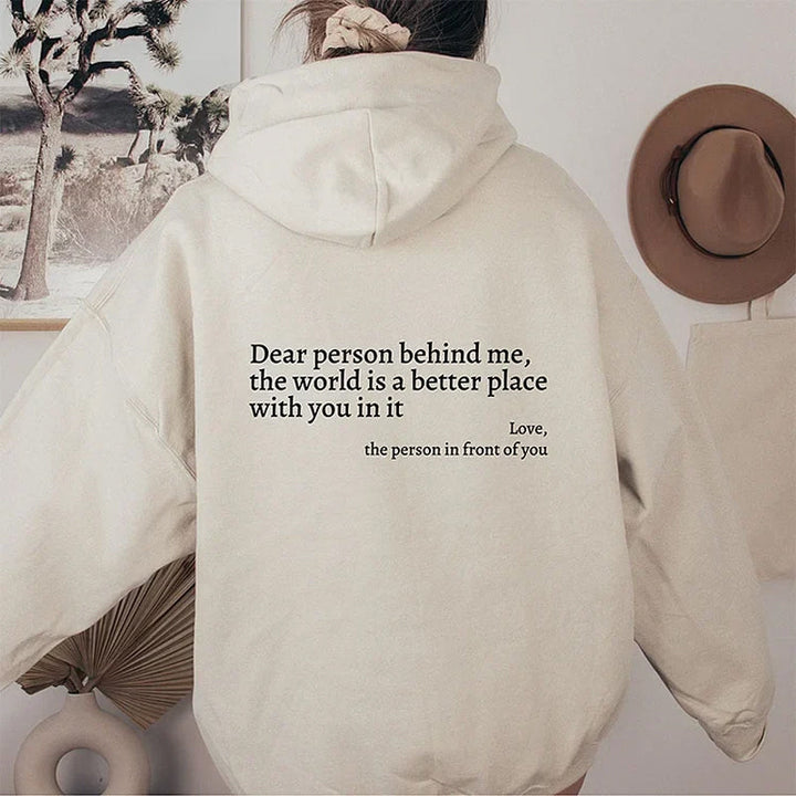 Frau in beigem Hoodie mit Aufschrift, Wanddekoration, Hut, Pflanzen im Hintergrund.