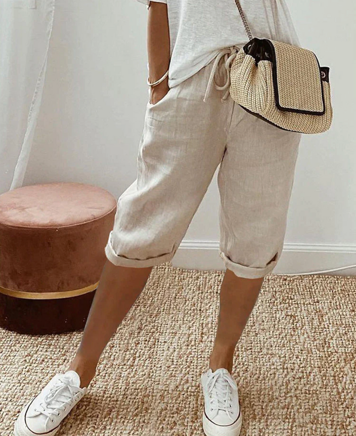 Frau in beigen Leinen-Shorts, weißem T-Shirt, weißen Sneakers, Strohtasche, lässiger Stil.