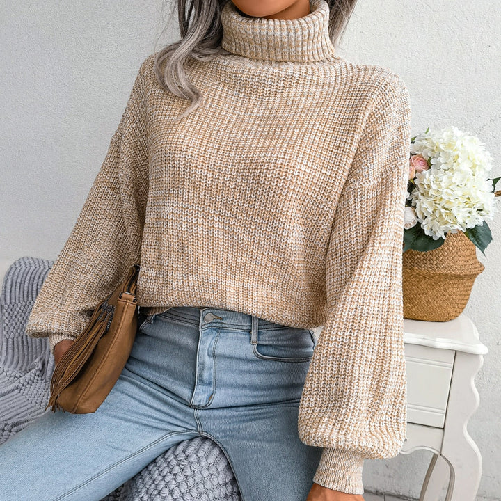 Frau in beigem Strickpullover mit Rollkragen, hellblauer Jeans, brauner Tasche, Blumen im Hintergrund.