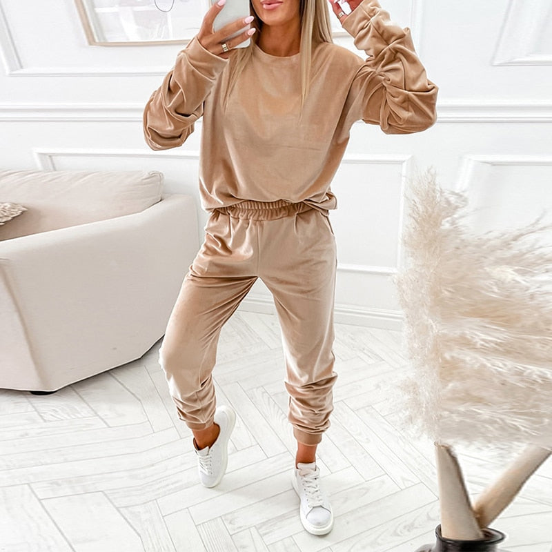 Frau in beigem Samt-Trainingsanzug, Wohnzimmer, weiße Sneakers, moderne Einrichtung.