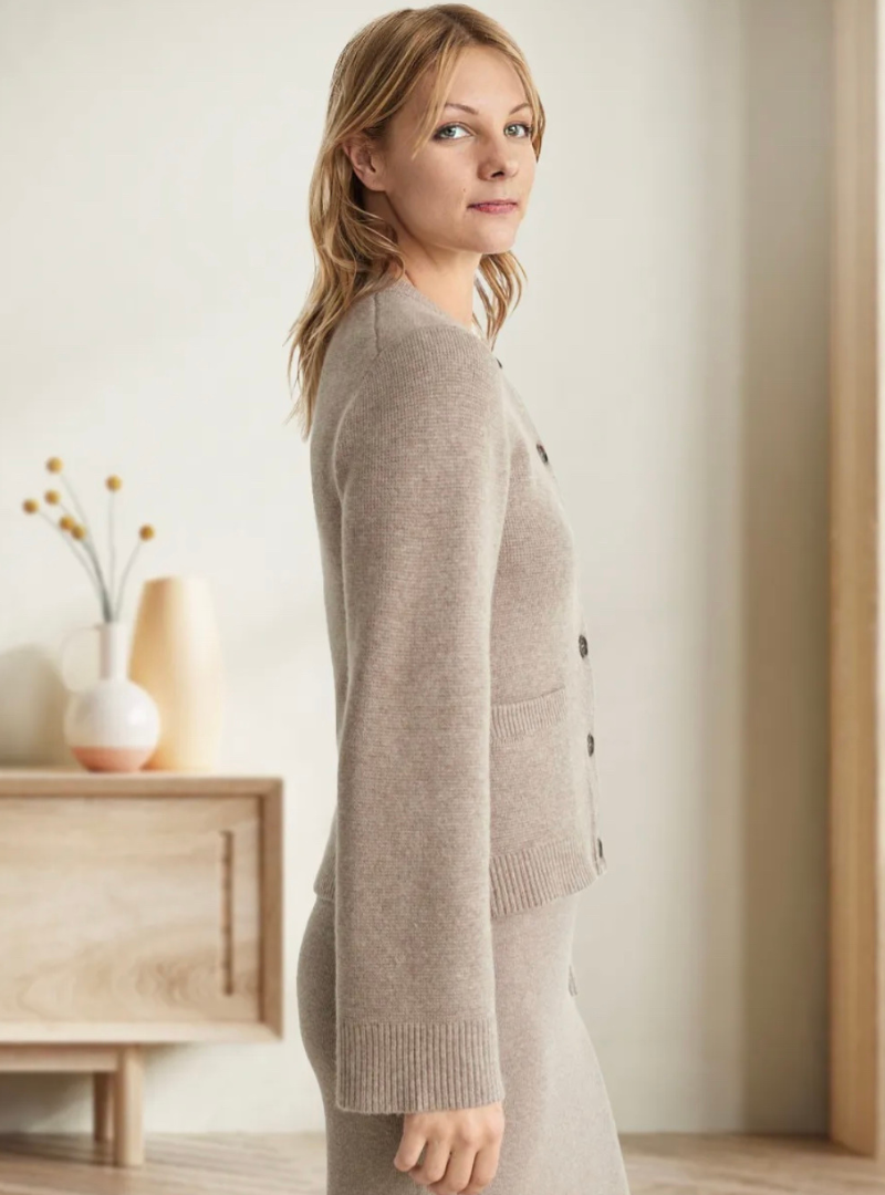 Frau in beigem Strickpullover, stehend vor Holzmöbeln, moderne Inneneinrichtung.