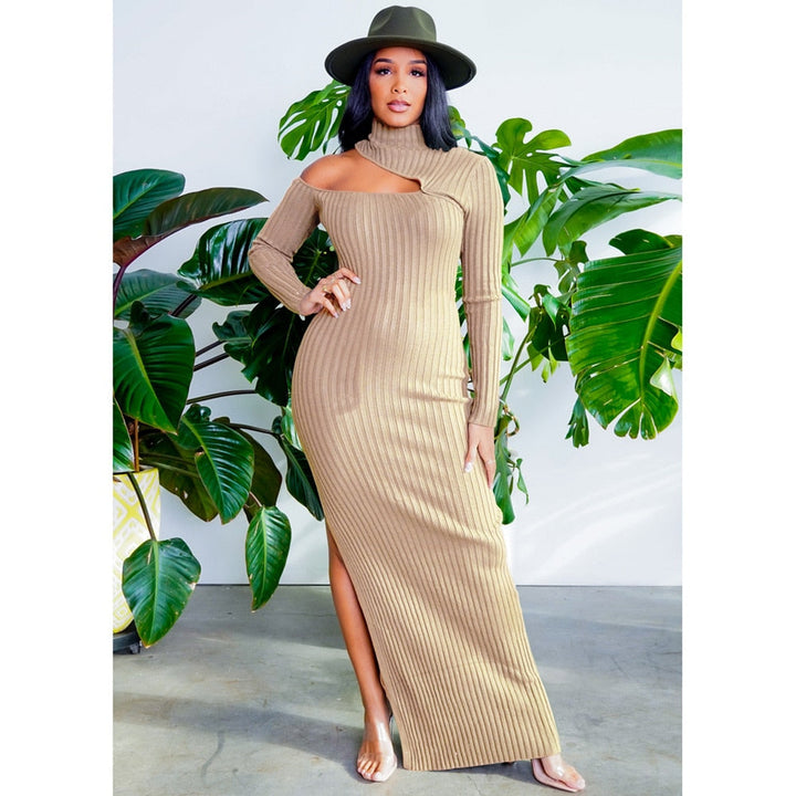 Frau in beige, schulterfreiem Strickkleid und Hut, vor grünen Pflanzen stehend.