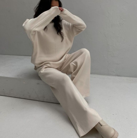 Frau in beigem Strickpullover und Hose, sitzt auf grauem Hintergrund, modisch, minimalistisch.