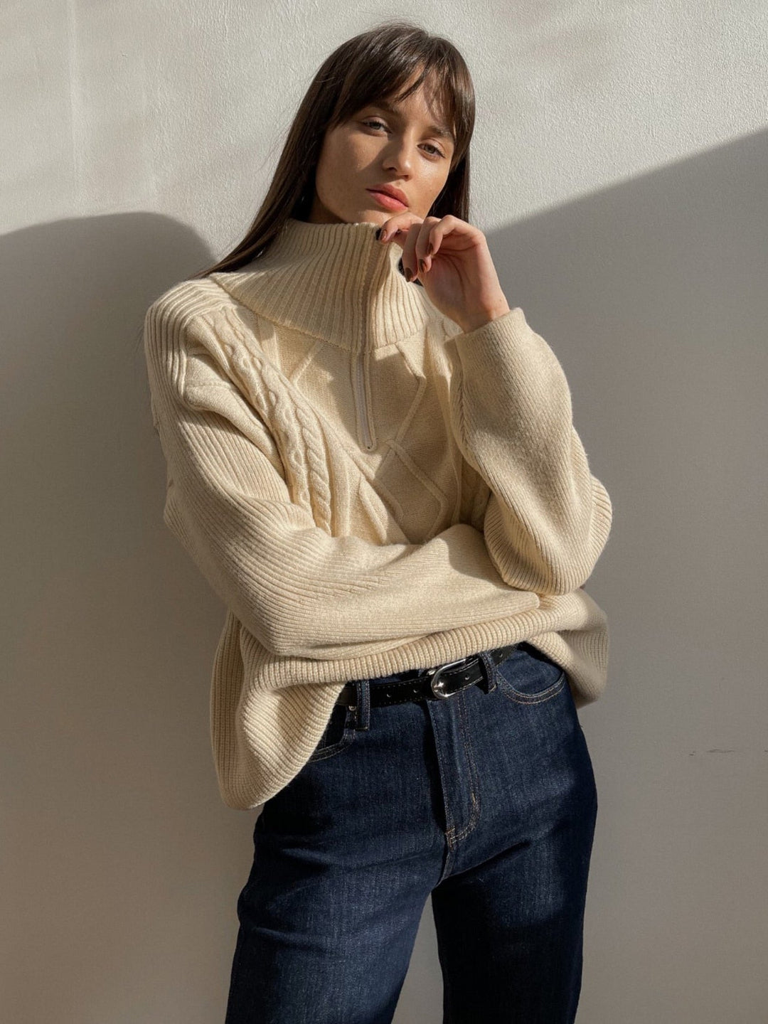 Frau in cremefarbenem Strickpullover mit Zopfmuster, Stehkragen, dunkle Jeans, lässiger Look.