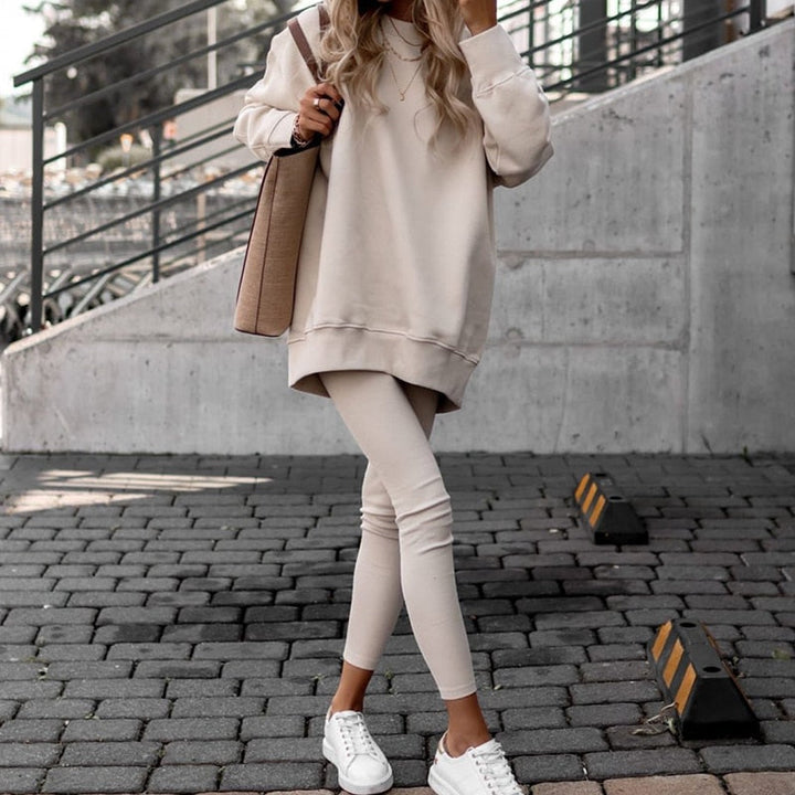 Frau in beigem Oversize-Pullover und Leggings, weiße Sneaker, beige Tasche, urbaner Hintergrund.